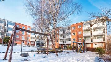 Un'area giochi con altalene e scivoli, circondata da terreno innevato e un edificio con numerose finestre.