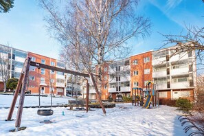 Un parque infantil con columpios y toboganes, rodeado de suelo cubierto de nieve y un edificio con numerosas ventanas.