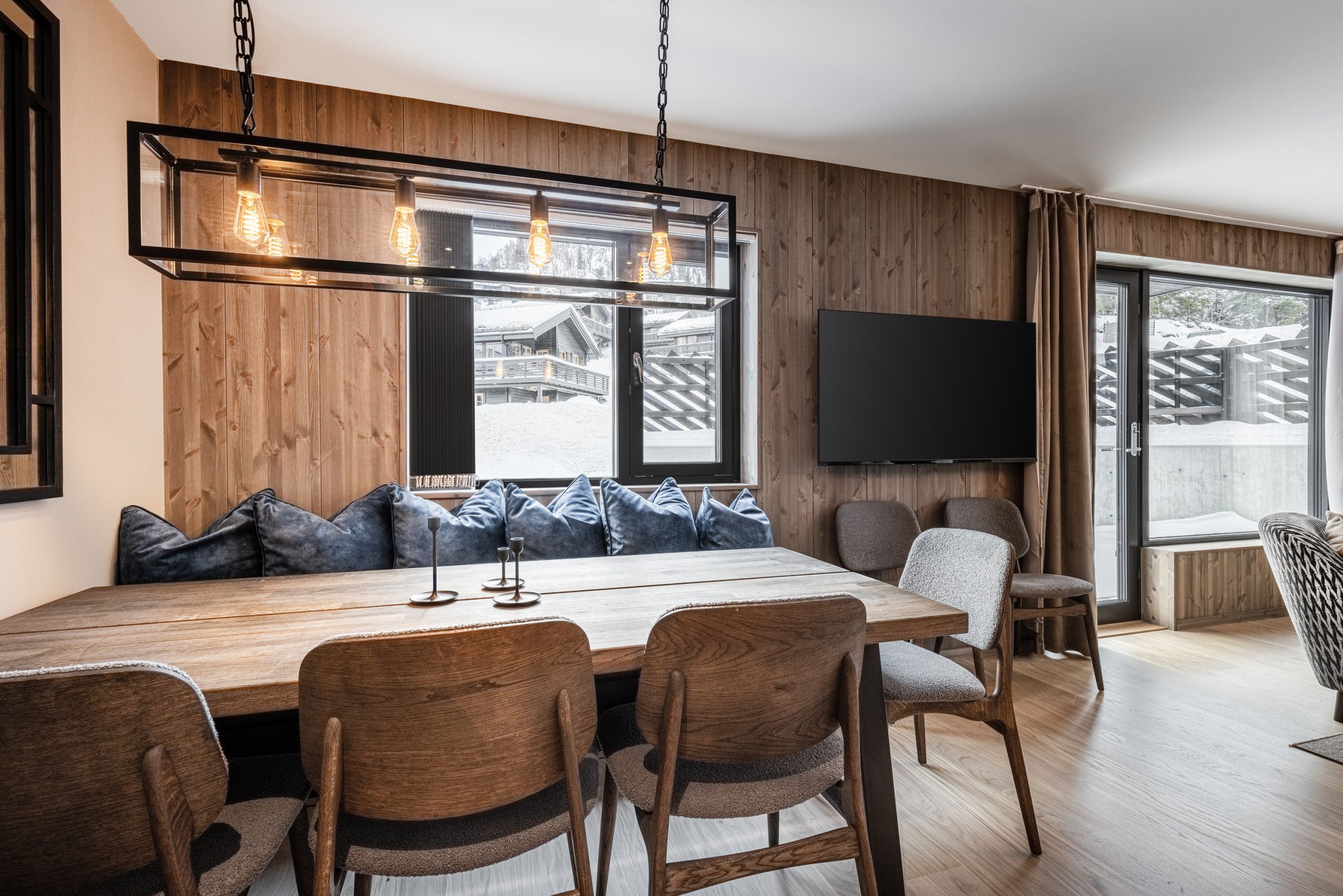Un comedor moderno con una mesa de madera, sillas tapizadas y un banco empotrado con cojines. Un televisor está montado en la pared, y la habitación tiene ventanas amplias con vista a un paisaje nevado.