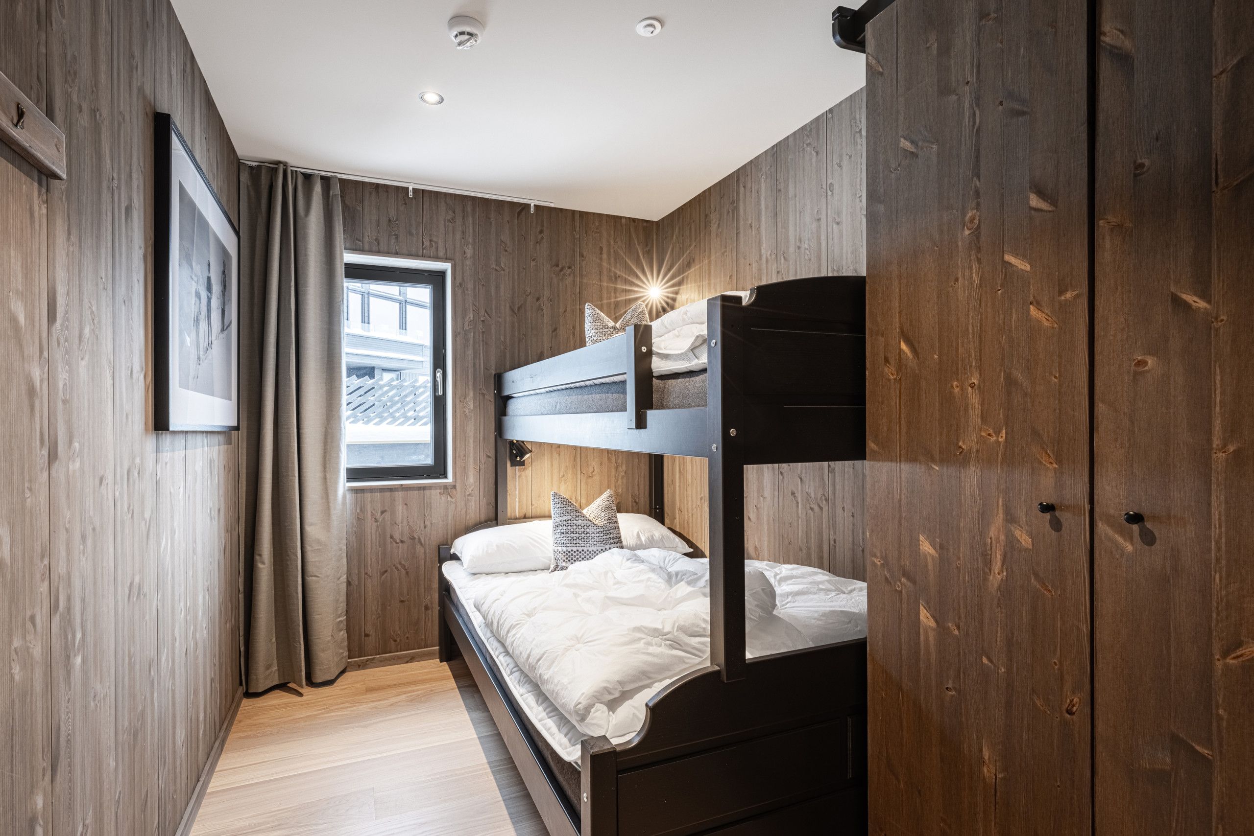 Een kamer met een stapelbed, houten wanden, een raam met gordijnen en een lijst met een foto aan de muur.