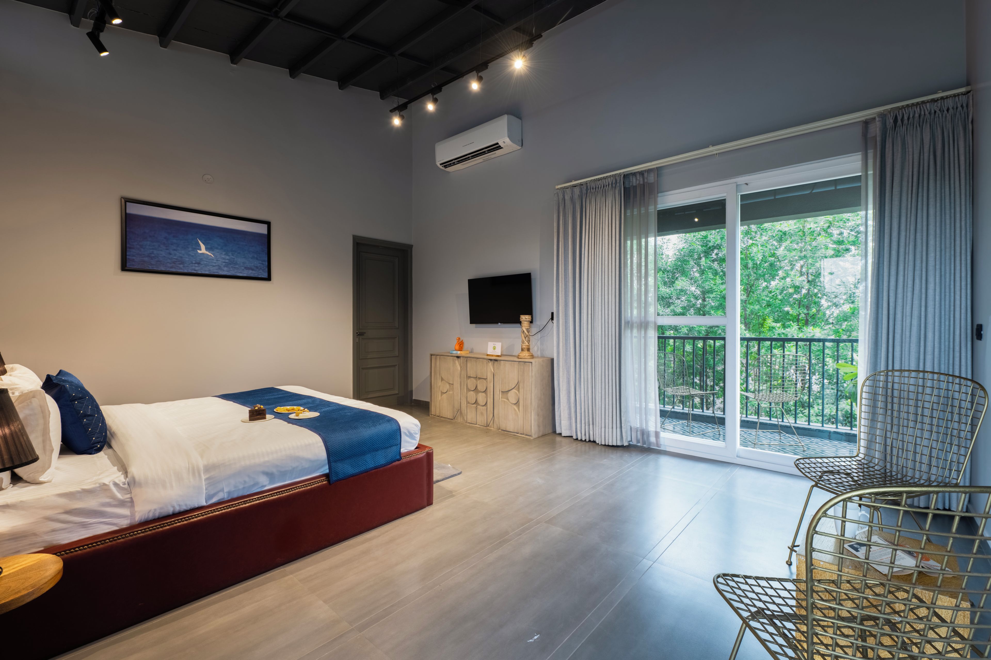A bedroom with a bed, a dresser, a television, a balcony with chairs, and a large window.