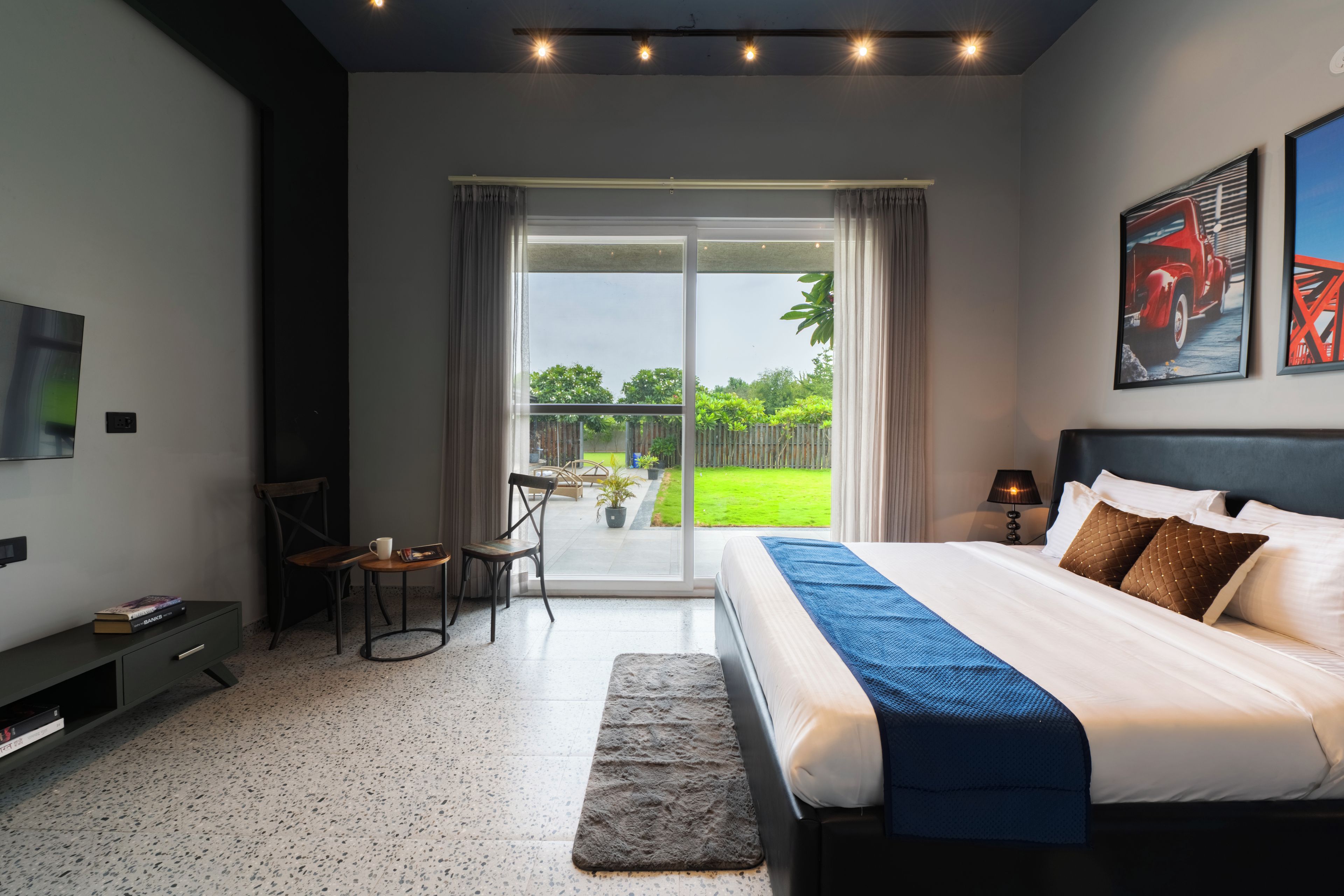 A modern bedroom with a large bed, a desk, and a chair. There is a sliding glass door leading to a balcony with a view of greenery.