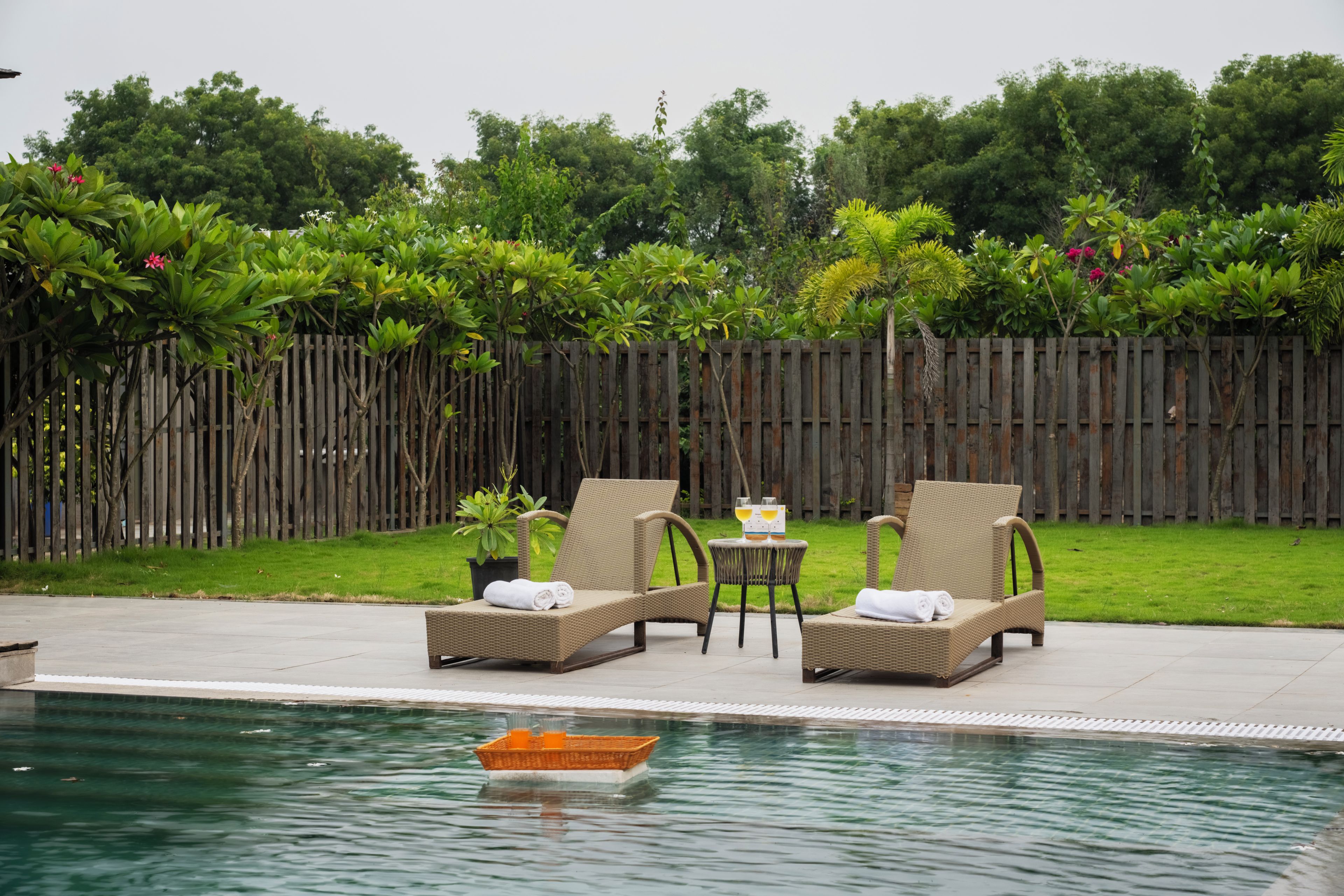 Zona junto a la piscina con dos sillones de mimbre, una mesita con dos vasos y una valla de madera al fondo.