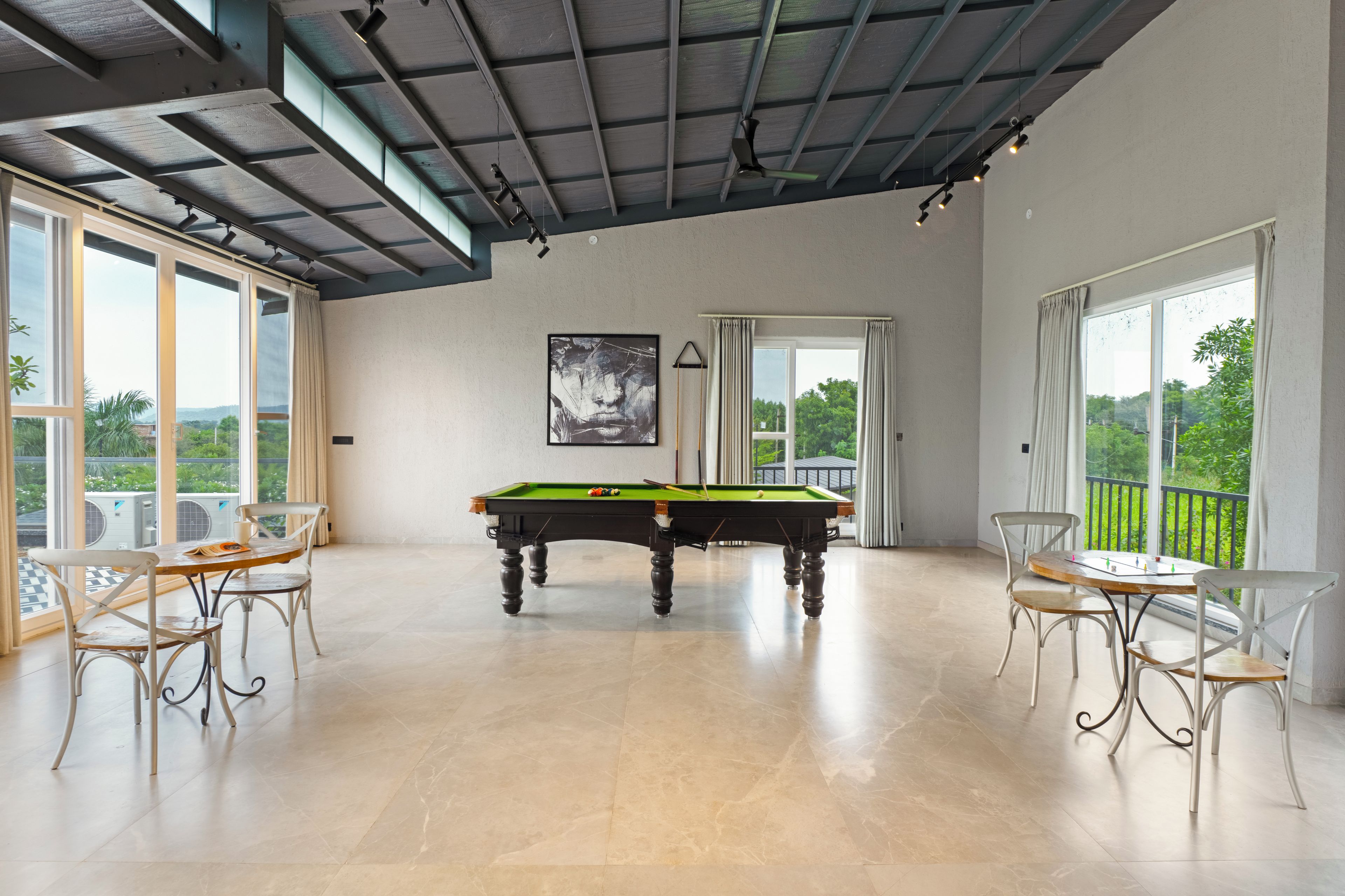 Une pièce spacieuse équipée d’une table de billard, de deux fauteuils et de grandes fenêtres donnant sur un paysage verdoyant.
