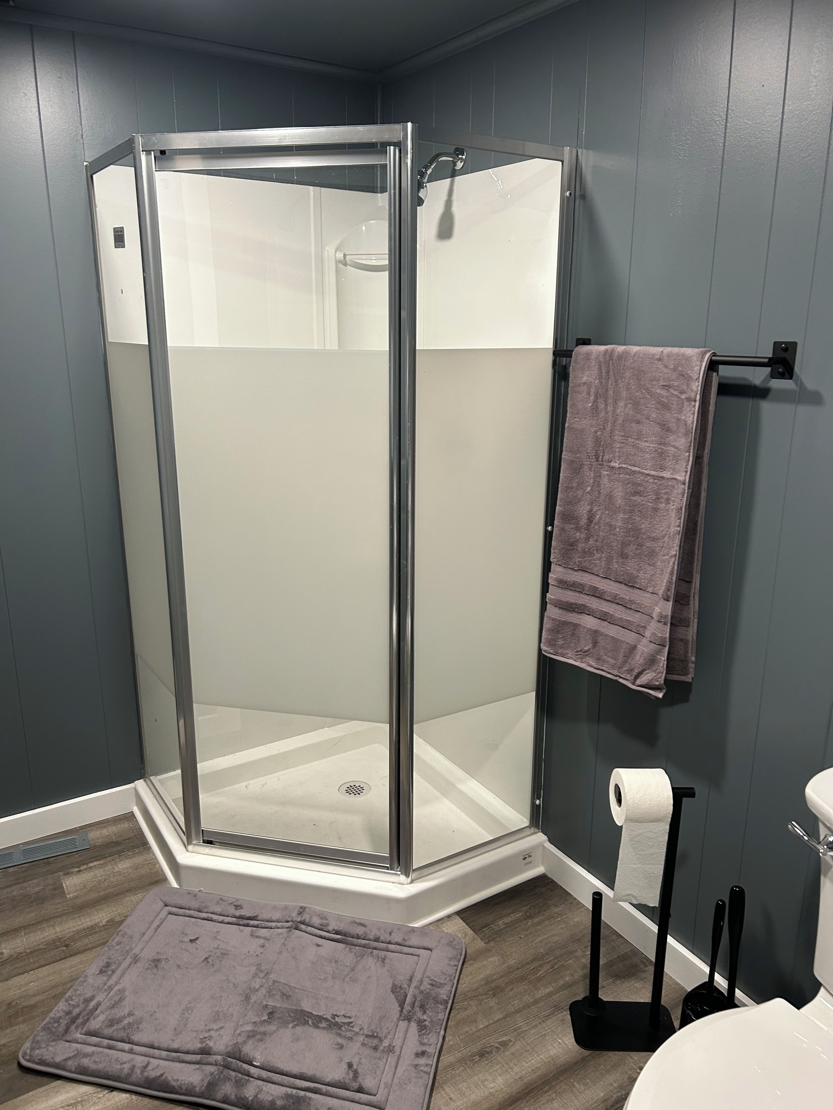 Cabina de ducha con puerta de vidrio, toallero con toalla, dispensador de papel higiénico y felpudo.