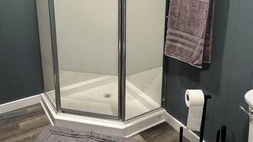 Cabina de ducha con puerta de vidrio, toallero con toalla, dispensador de papel higiénico y felpudo.