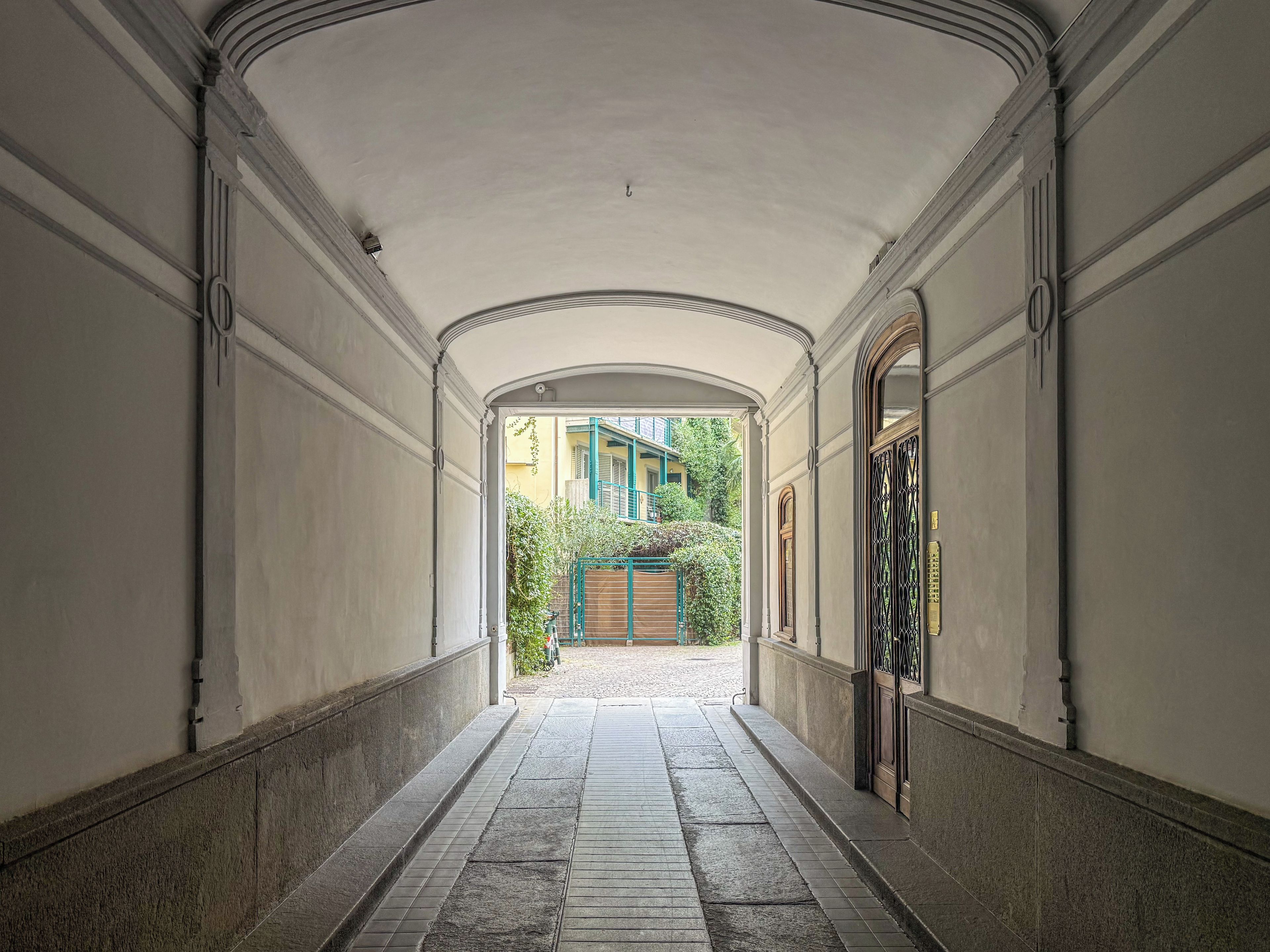 A corridor with a tiled floor, arched ceiling, and a view of a building with a green gate and steps.