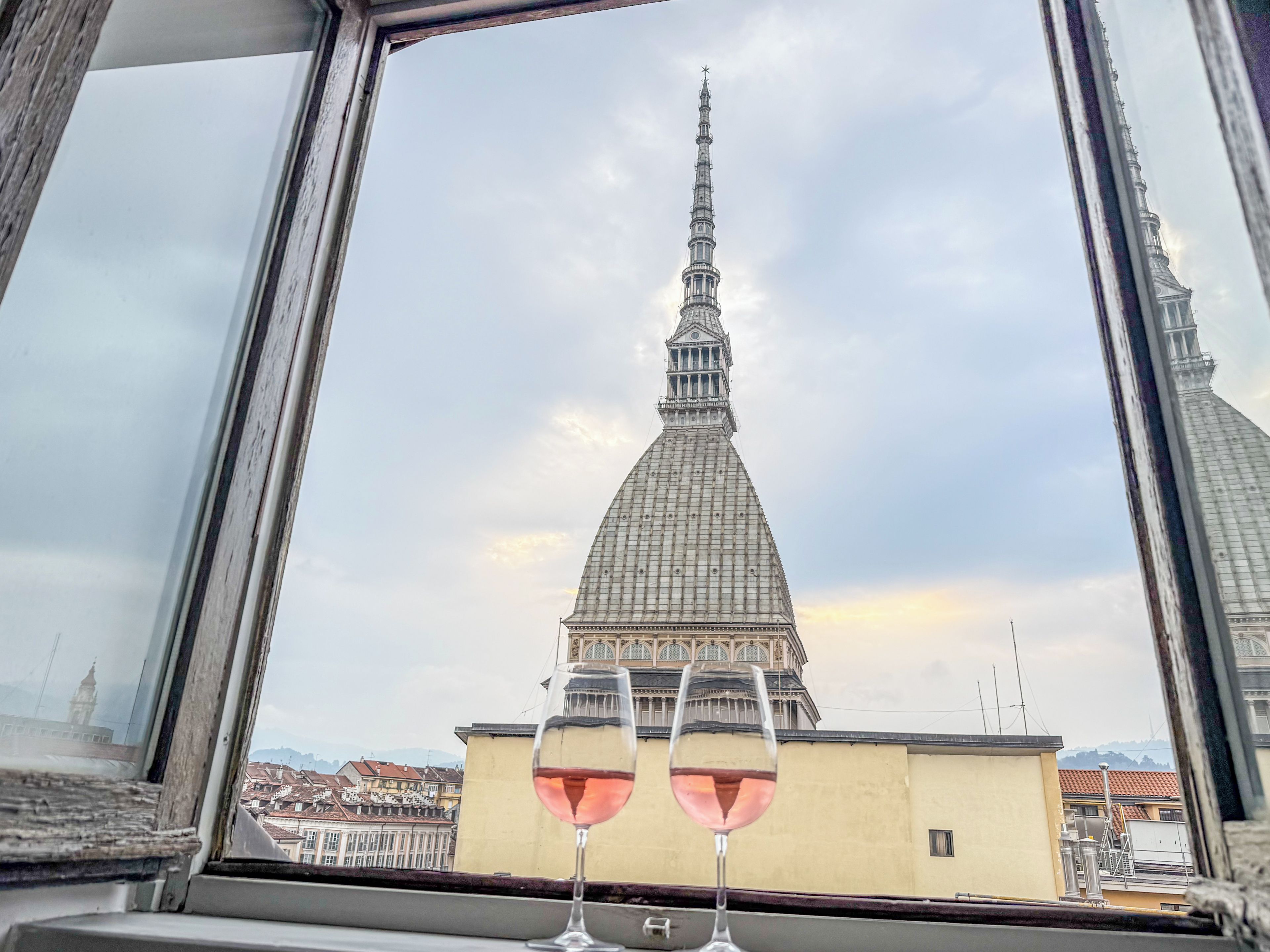 Vista desde una ventana de un edificio con una torre destacada, dos copas de vino tinto sobre el alféizar y un cielo despejado.