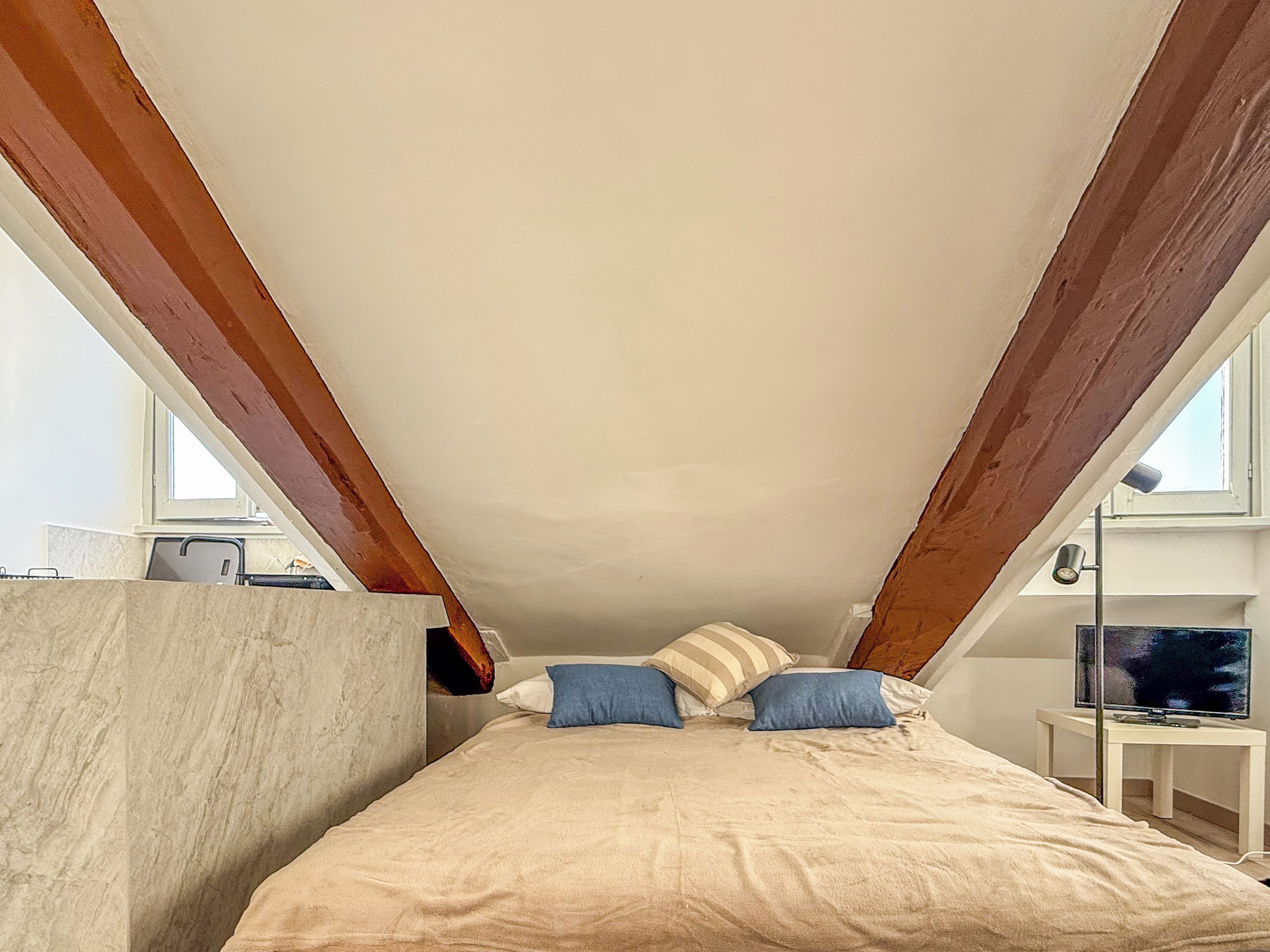 Un dormitorio con techo inclinado, una cama con cubierta color beige, dos almohadas, una mesita de noche con una lámpara y un televisor sobre un soporte.