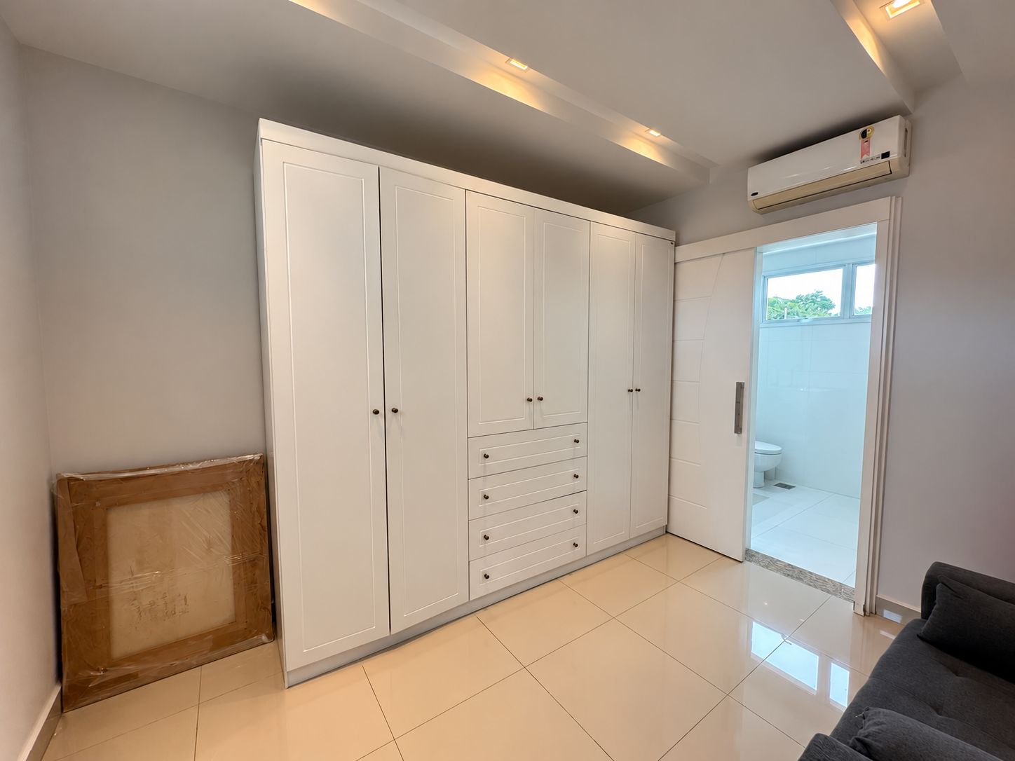 Quarto com um guarda-roupa branco grande, um sofá cinza e uma porta de vidro deslizante que dá para uma varanda.