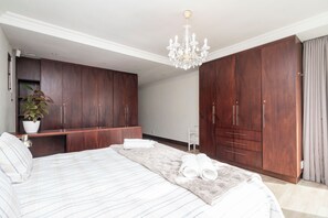 A bedroom with a large bed, a chandelier, wooden wardrobes, and a potted plant. - 5BR Sea Point Oasis: Main House + Flatlet - Solar Powered - Heated Pool & Garage (Cape Town)