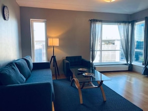 Una sala moderna con sofá, una mesita de centro y una lámpara. Se ve una ventana con cortinas y vistas a la ciudad.