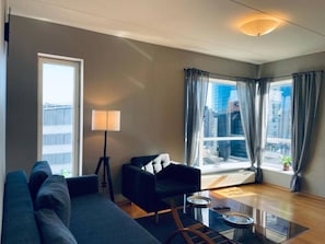 Un salon moderne avec un canapé, un fauteuil noir, une table basse en verre et un lampadaire. Une grande fenêtre, ornée de rideaux, offre une vue sur la ville.