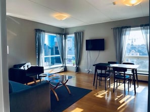 Un salon moderne comprenant un canapé, une table basse, une table à manger avec des chaises, un téléviseur à écran plat fixé au mur et de grandes fenêtres avec des rideaux.