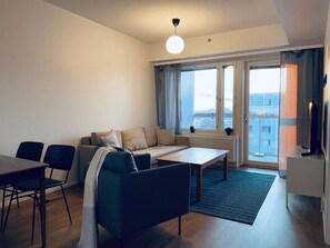 Un salon moderne avec un canapé, une table basse et des fauteuils. On y trouve une grande fenêtre avec des rideaux, un radiateur et un téléviseur posé sur un meuble.