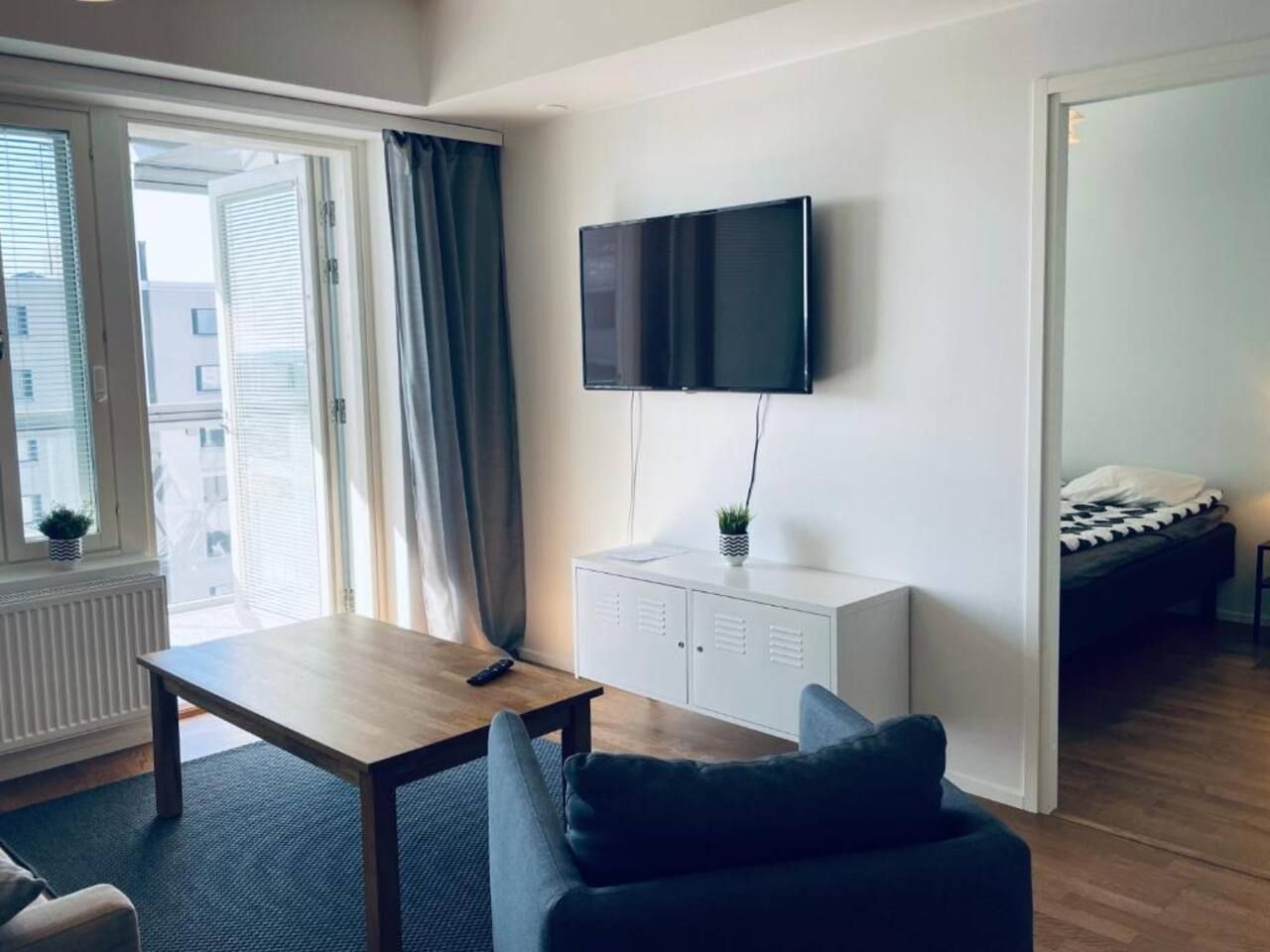 A modern living room with a flat-screen TV mounted on the wall, a wooden coffee table, a dark sofa, and a bed in the adjacent room.