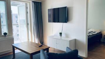 A modern living room with a flat-screen TV mounted on the wall, a wooden coffee table, a dark sofa, and a bed in the adjacent room.