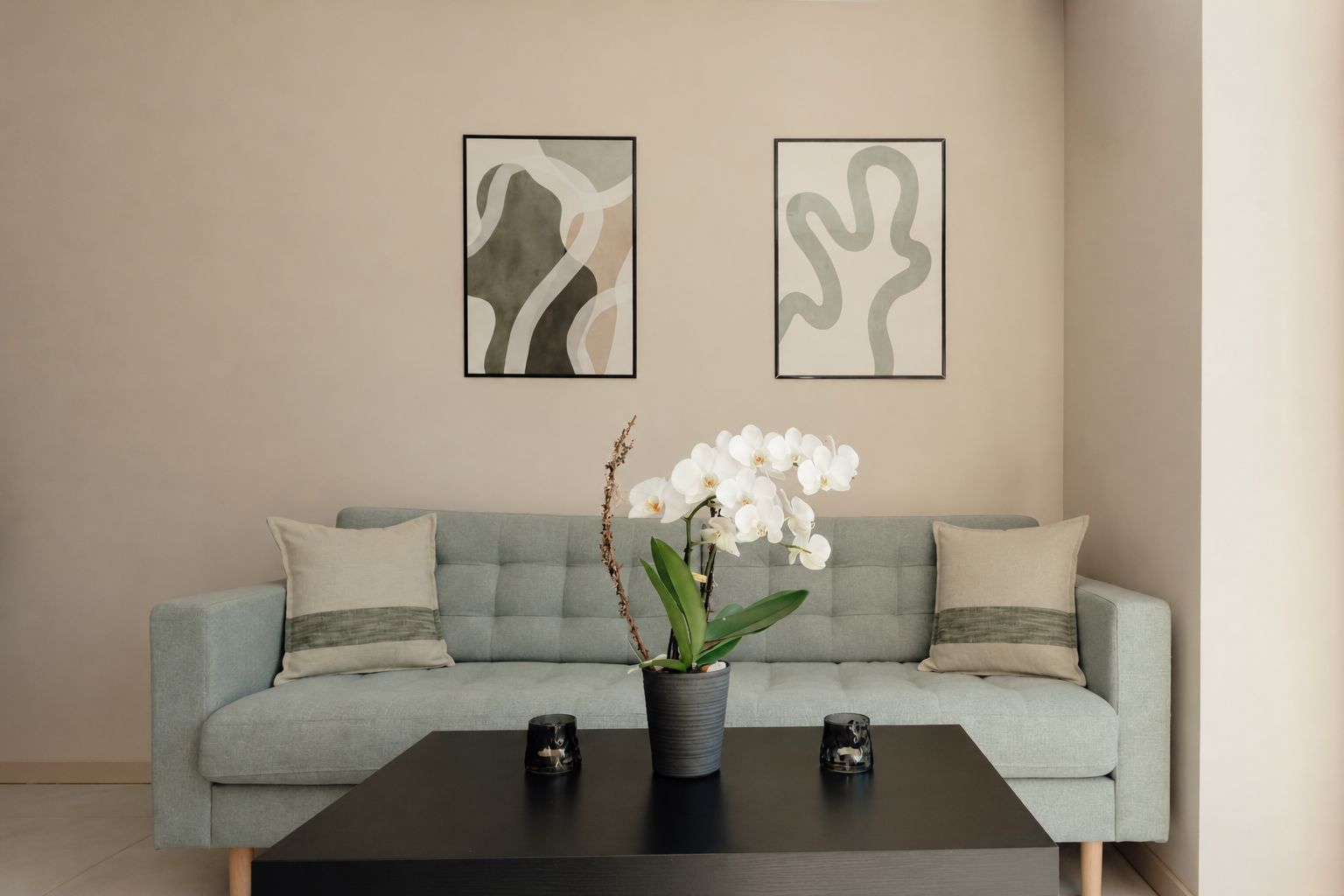 A modern living room with a grey sofa, two framed abstract artworks, a potted orchid, and two candles on a dark coffee table.