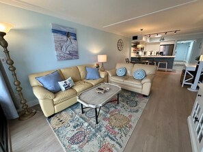 Sala de estar com dois sofás, uma mesa de centro e um tapete. Há um quadro na parede e uma cozinha ao fundo.