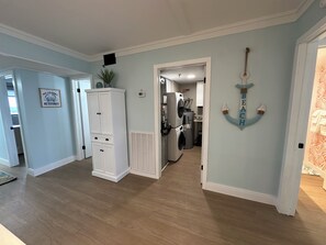 A hallway with a white cabinet, a wall-mounted air vent, and a door leading to a kitchen area with a refrigerator and washing machine. - Sleeps 6 • Oceanfront • Panoramic Views • Private Balcony • Indoor & Outdoor Pools • Hot Tub • Elevator • Direct Beach Access • Washer & Dryer (Carolina Beach)