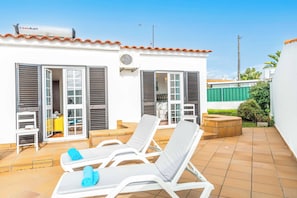 Ein Wohnhaus mit bepflasterter Terrasse, weißen Wänden und einem roten Ziegeldach. Zwei offene Türen führen in einen Raum mit einem Bett und einem kleinen Tisch.