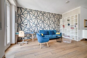 A modern living room with a geometric patterned wall, a blue sofa, a wooden floor, and a white shelving unit. - Hostmate - Milano - C. Buenos Aires - City Center (Milan)