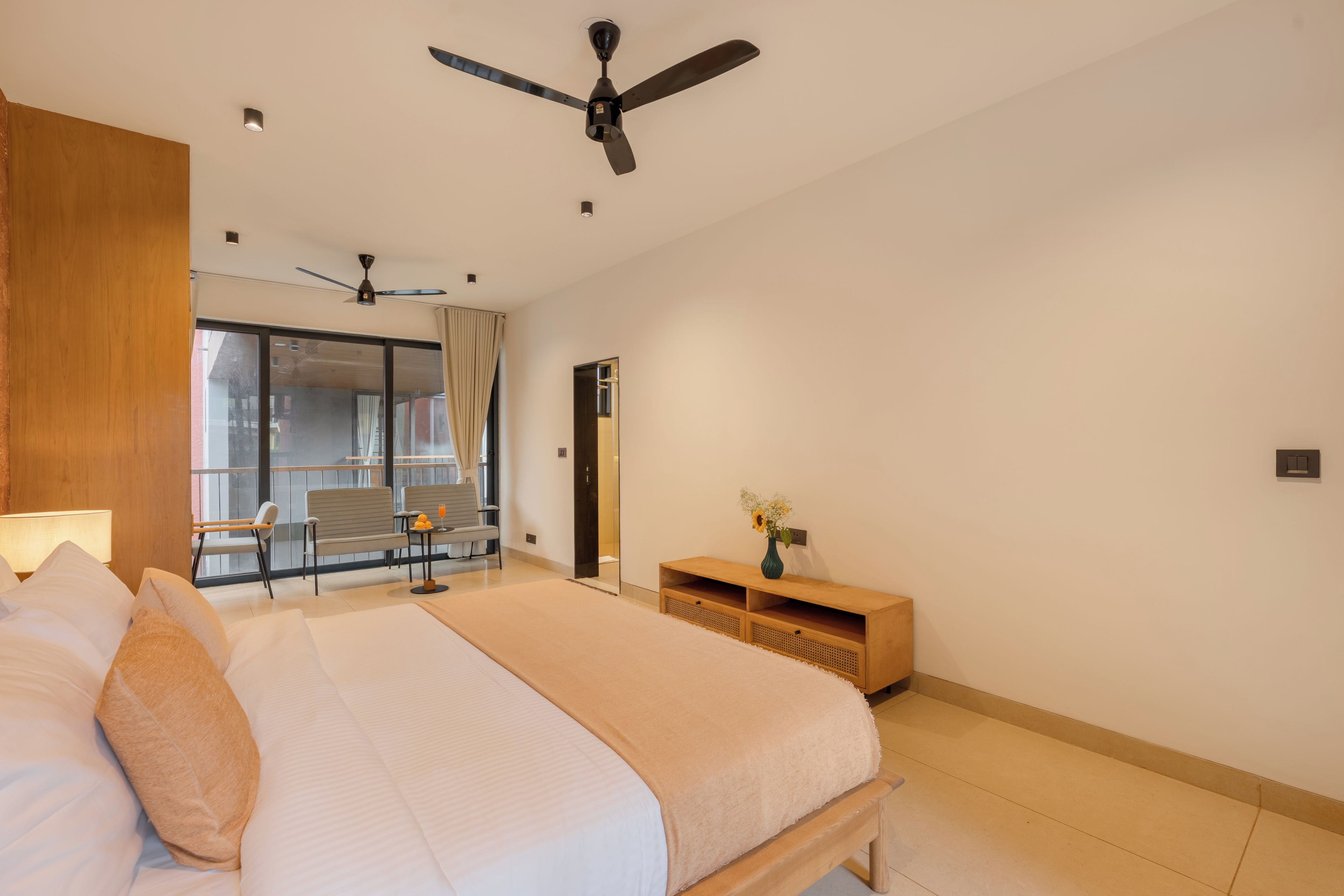 Ein modernes Hotelzimmer mit einem großen Bett, einem Deckenventilator, einer Sitzecke mit Tisch und Stühlen sowie einem Ausblick durch ein großes Fenster.