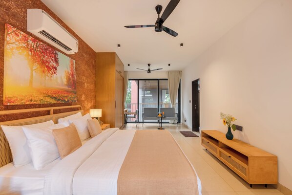 Une chambre d’hôtel avec un grand lit, un ventilateur de plafond, un tableau représentant une forêt, un bureau avec une chaise et une vue sur l’extérieur.