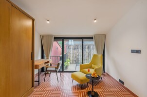 Una habitación moderna con un armario de madera, un escritorio, un sillón amarillo y vistas a un edificio rojo a través de una puerta de cristal.
