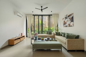 A modern living room with a sofa, ottoman, and a coffee table. There is a large window with curtains, a ceiling fan, and a wall-mounted air conditioner. - ELIVAAS Signature Coco Baia 3BHK Villa (Marra)