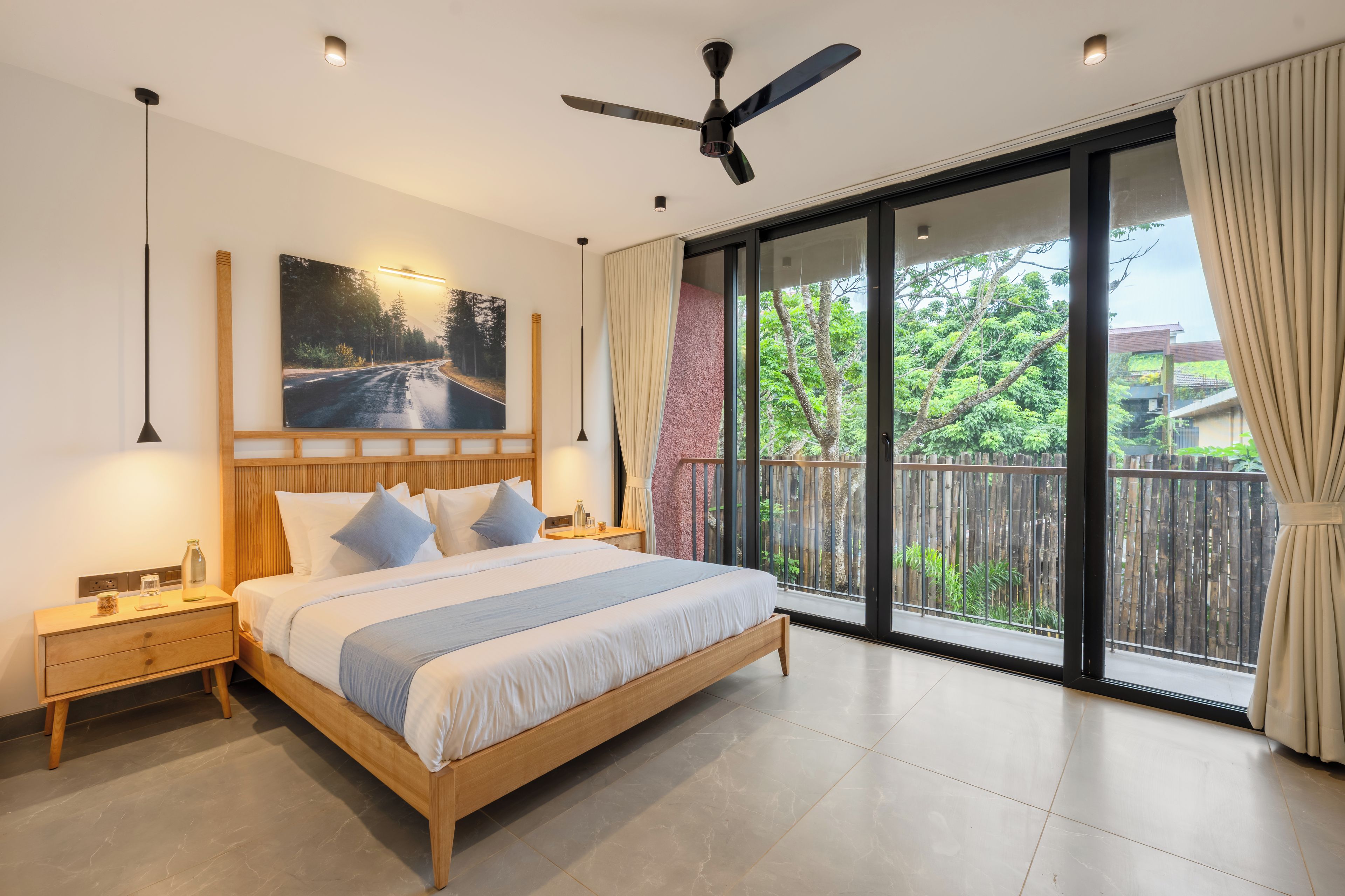 A bedroom with a large bed, a ceiling fan, a nightstand, a bedside table, a painting, and a view of greenery through a sliding glass door.