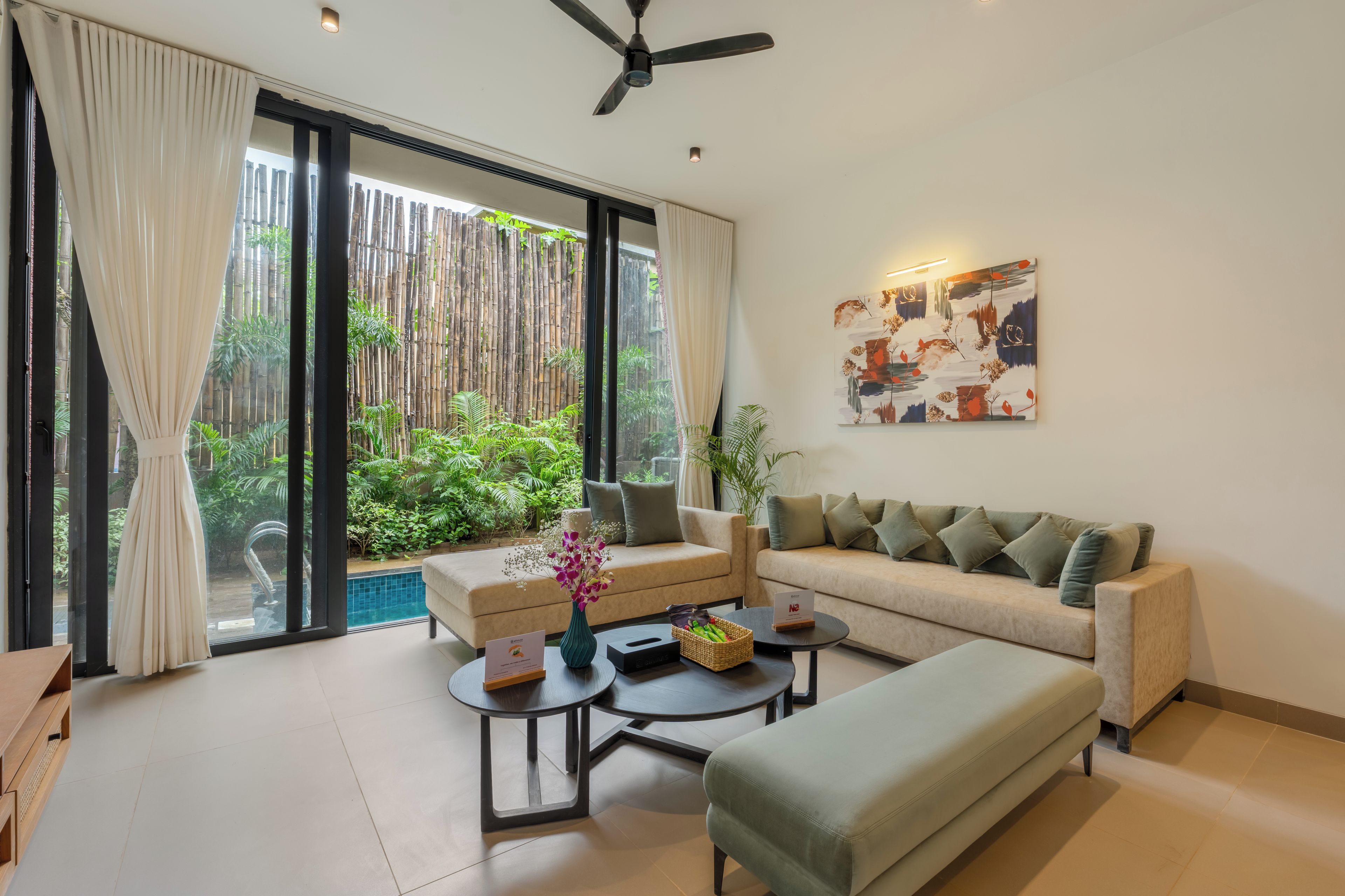 A modern living room with a sofa, two armchairs, a coffee table, and a view of a bamboo fence and greenery outside.