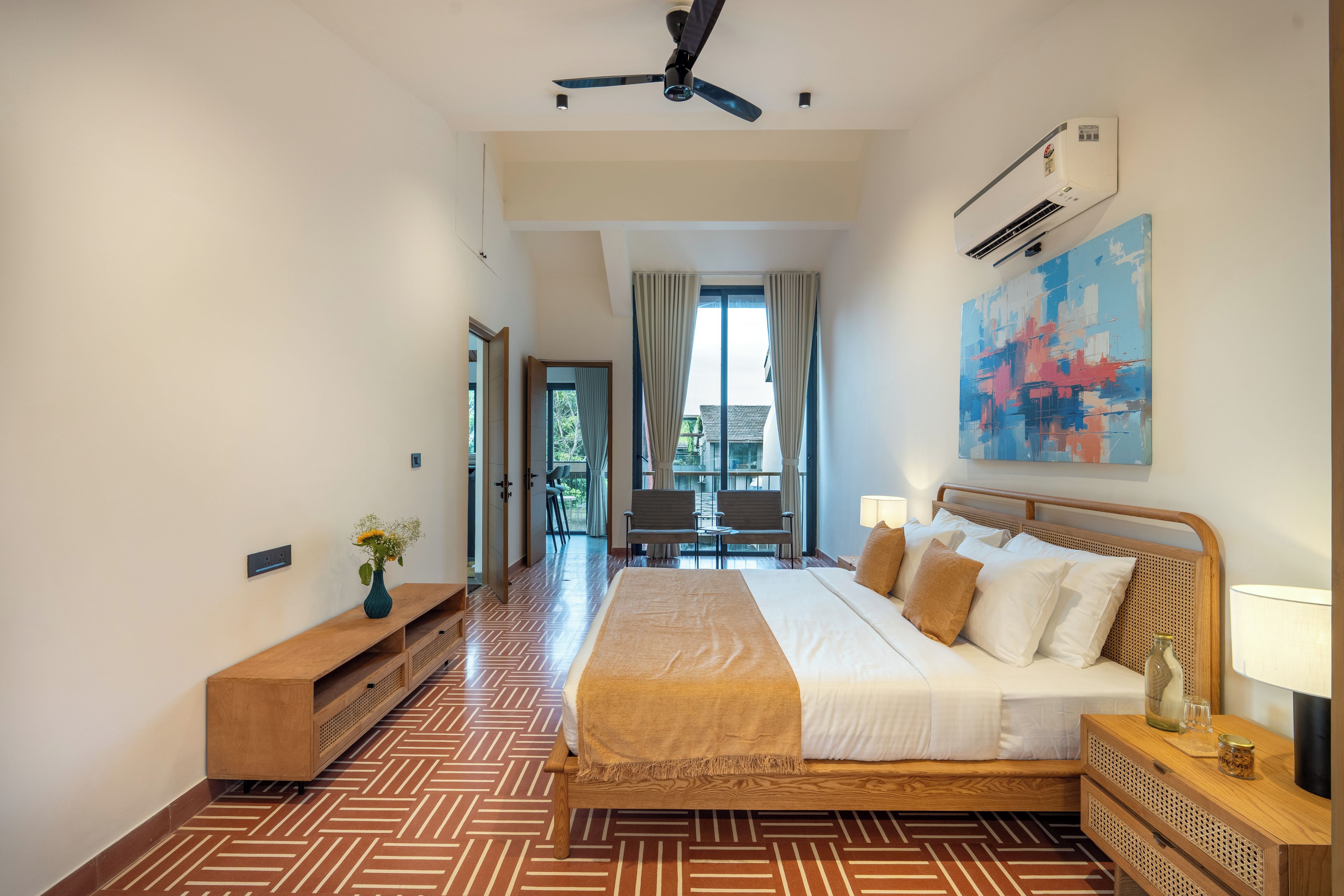 A hotel room with a large bed, a ceiling fan, an air conditioning unit, a painting on the wall, and a view of the outside.