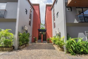 Edifício com entrada central ladeada por dois pilares de tijolos vermelhos, pátio pavimentado e plantas em vasos.