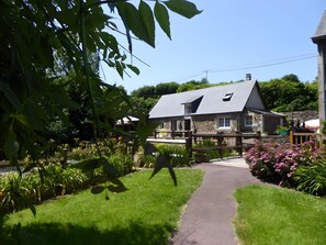 Ein Gebäude aus Stein mit einem geneigten Dach, einem gepflasterten Weg zum Eingang und einem Garten mit verschiedenen Pflanzen und Blumen.