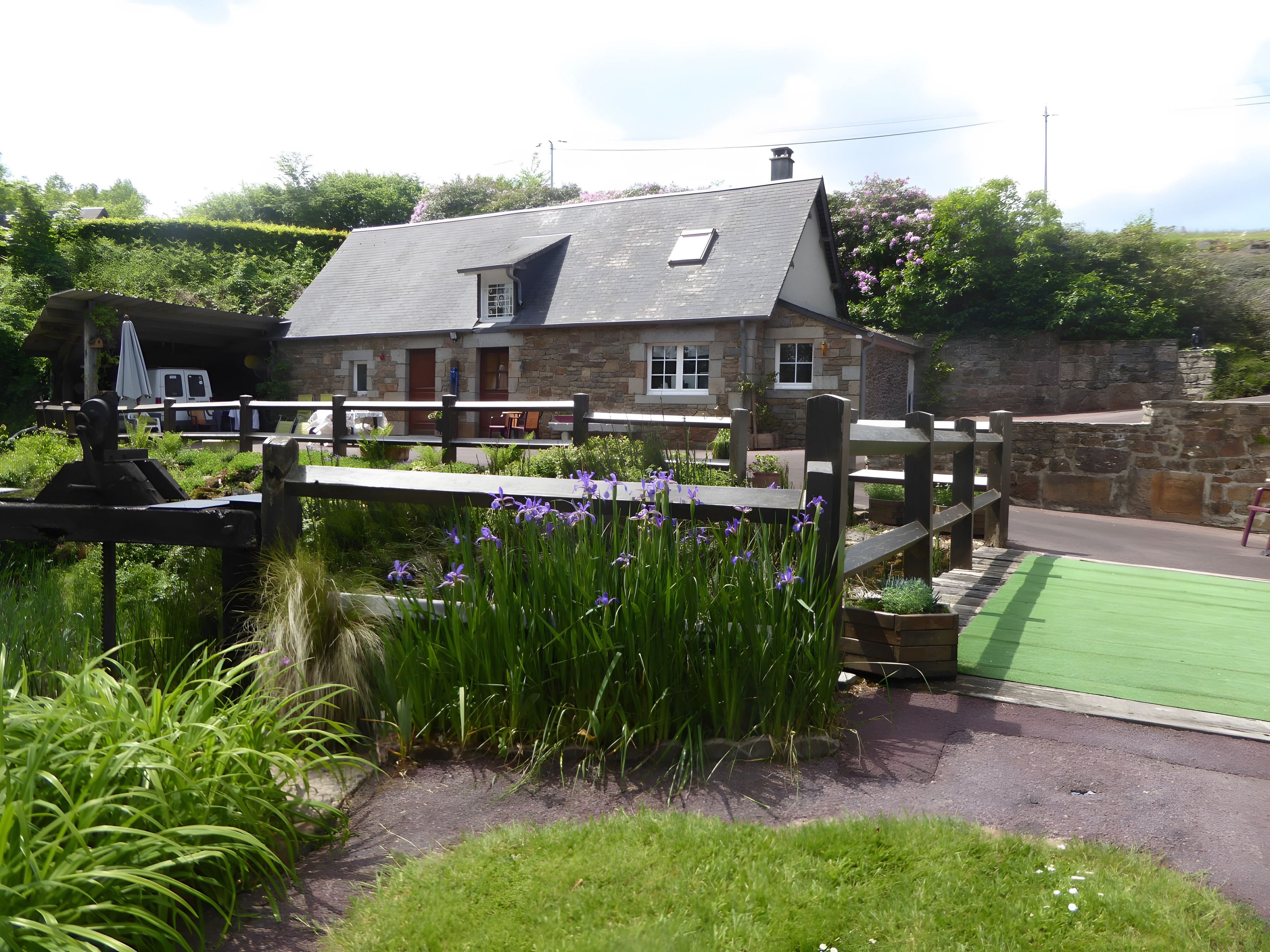 A stone building with a sloped roof, a fenced garden area with purple flowers, and a paved path.