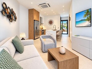 Ein modernes Wohnzimmer mit Sofa, Sessel und Couchtisch, eine Küche mit Kühlschrank und Herd sowie ein Essbereich mit Stühlen.