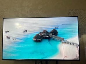 Una pantalla grande muestra una vista aérea de bungalows sobre el agua unidos por un puente, con barcos y tranquilas aguas turquesas.
