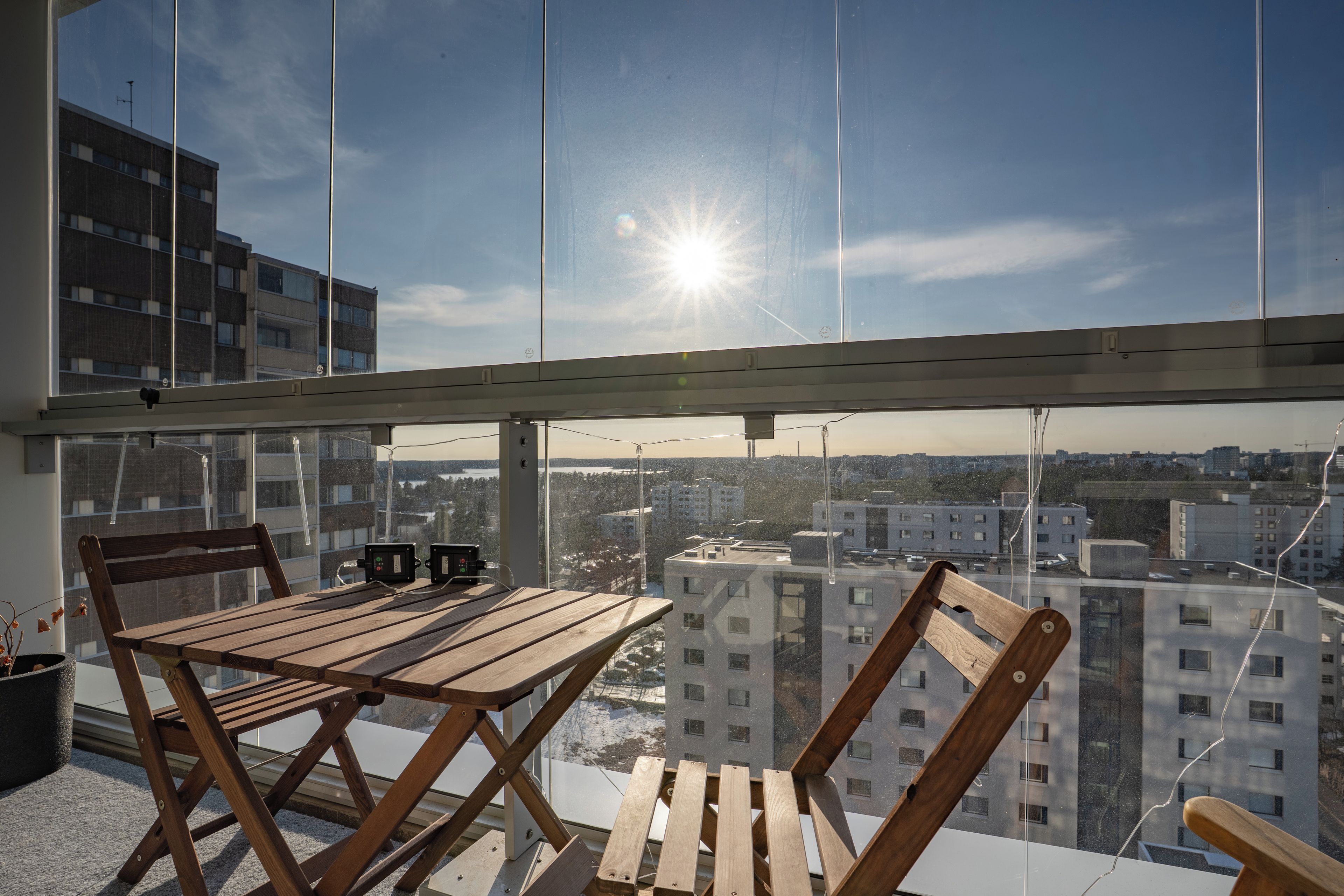 A balcony with a wooden table and chairs, a potted plant, and a view of a cityscape.