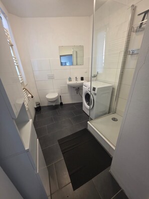 A modern bathroom with a glass shower enclosure, a toilet, a sink, and a washing machine. - Cozy apartment in the KTV with a terrace (Rostock)