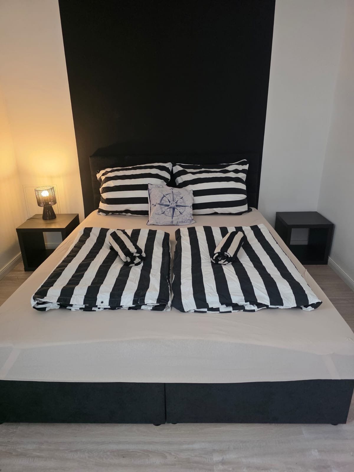 Una cama con ropa de cama a rayas blancas y negras, dos almohadas y una lámpara de noche. También hay una almohada con motivos de brújula.