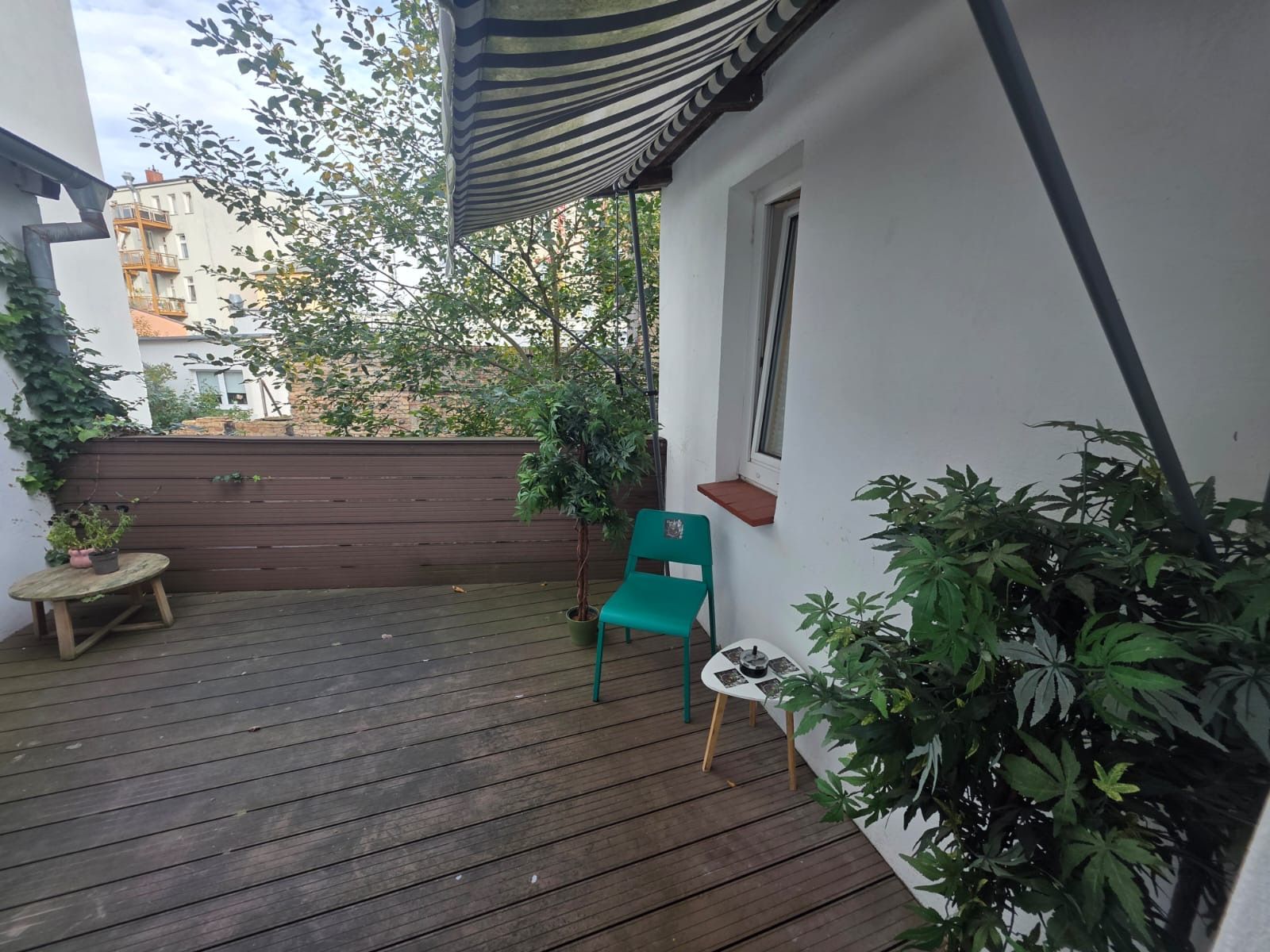 A wooden deck with a green chair and a small table, surrounded by potted plants and a striped awning.