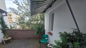 A wooden deck with a green chair and a small table, surrounded by potted plants and a striped awning.