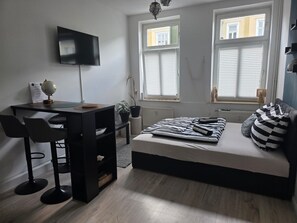 A bedroom with a bed, a desk with a chair, a television, and two windows with blinds.
