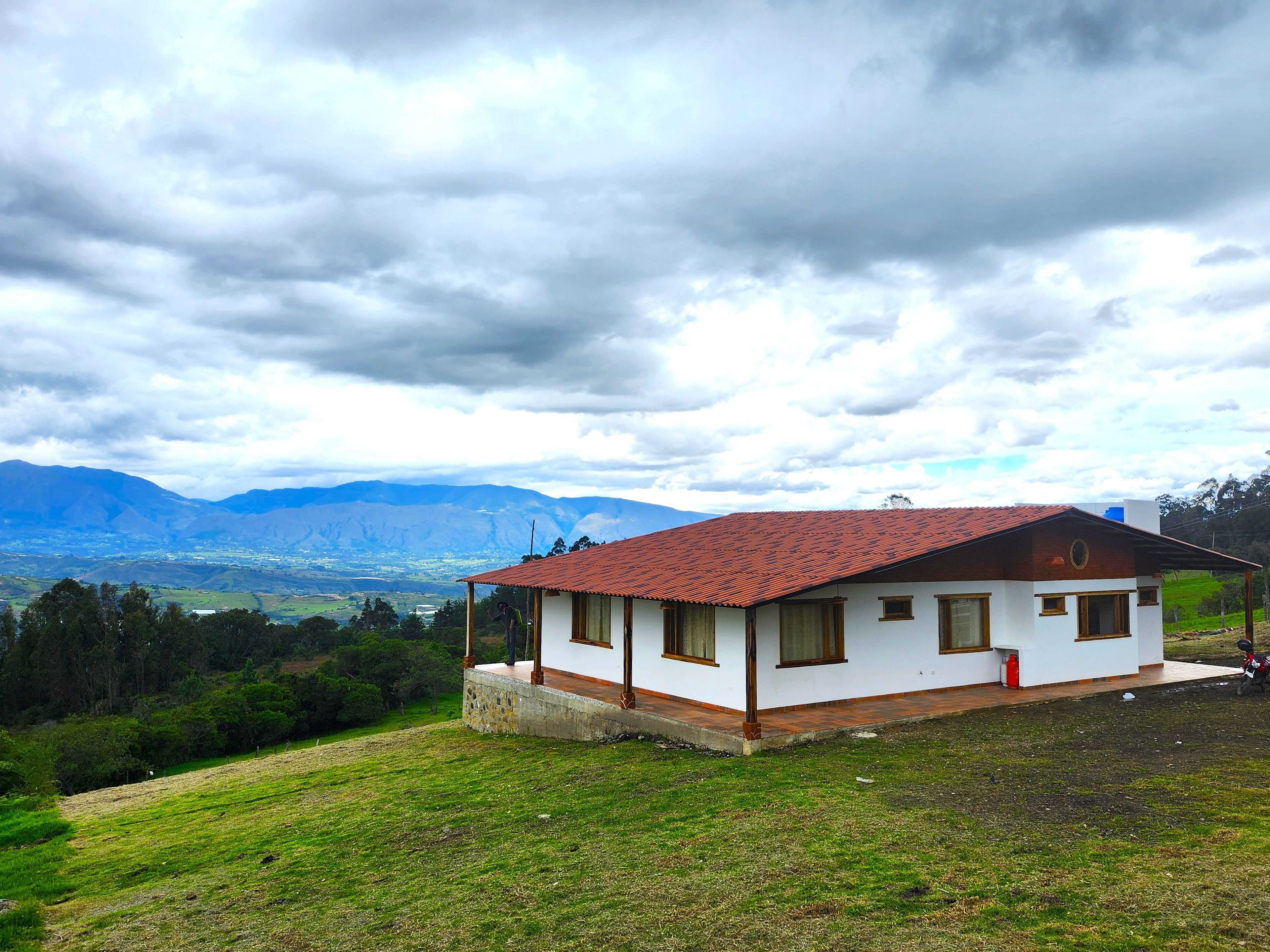 A single-story house with a red-tiled roof, surrounded by a grassy landscape and mountains in the background.