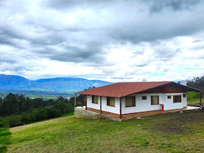 A single-story house with a red-tiled roof, surrounded by a grassy landscape and mountains in the background.