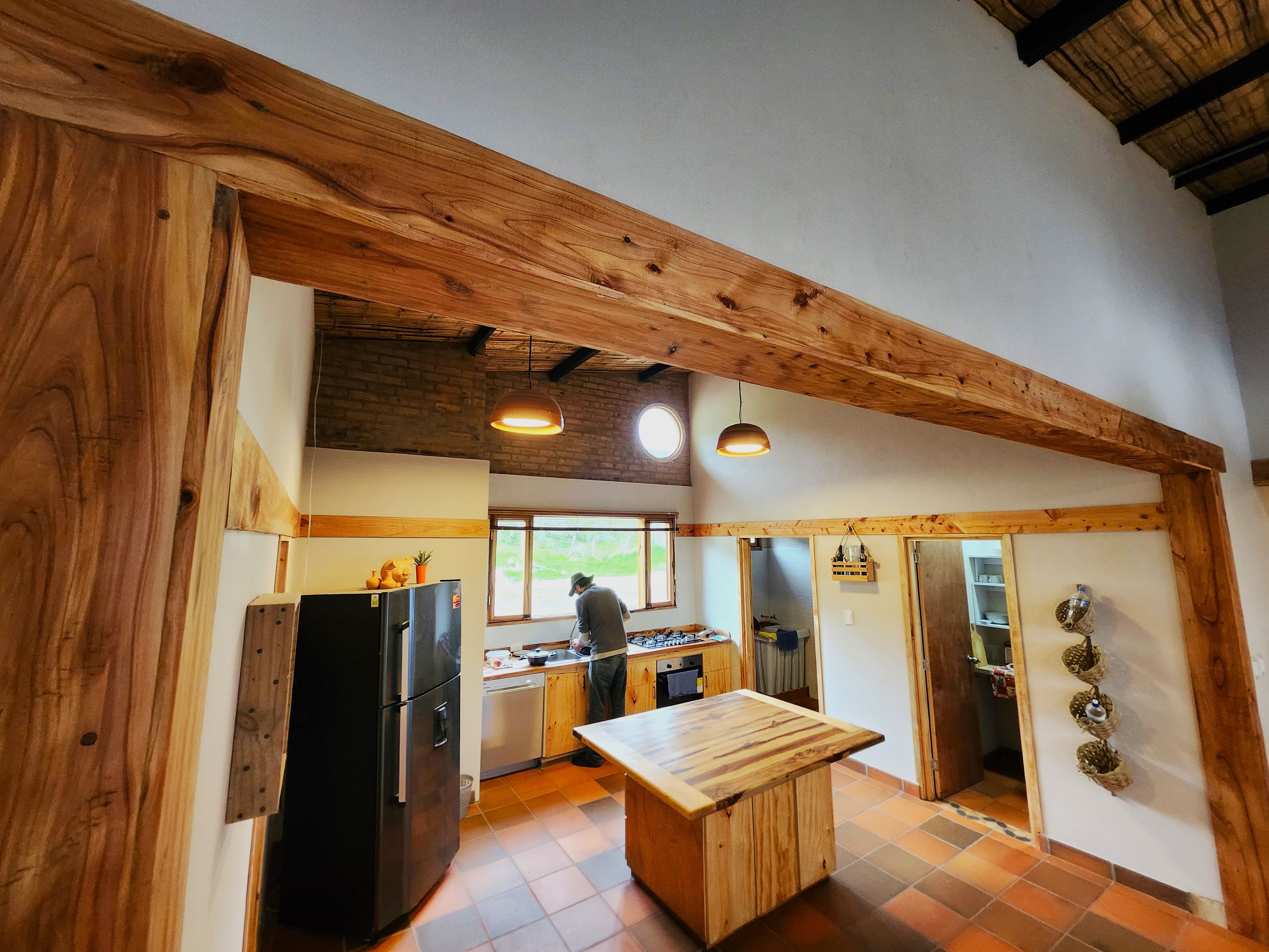 A kitchen with wooden beams, a black refrigerator, a wooden table, and a person cooking.