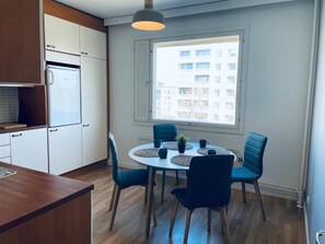 A compact kitchen with white cabinets, a built-in refrigerator, a round dining table with blue chairs, and a window with a view of buildings. - Spacious 1BR Near Metro and Major Shopping Center (Espoo)