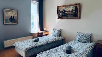 A room with two beds, a window, a radiator, a desk, a lamp, and two paintings on the wall.