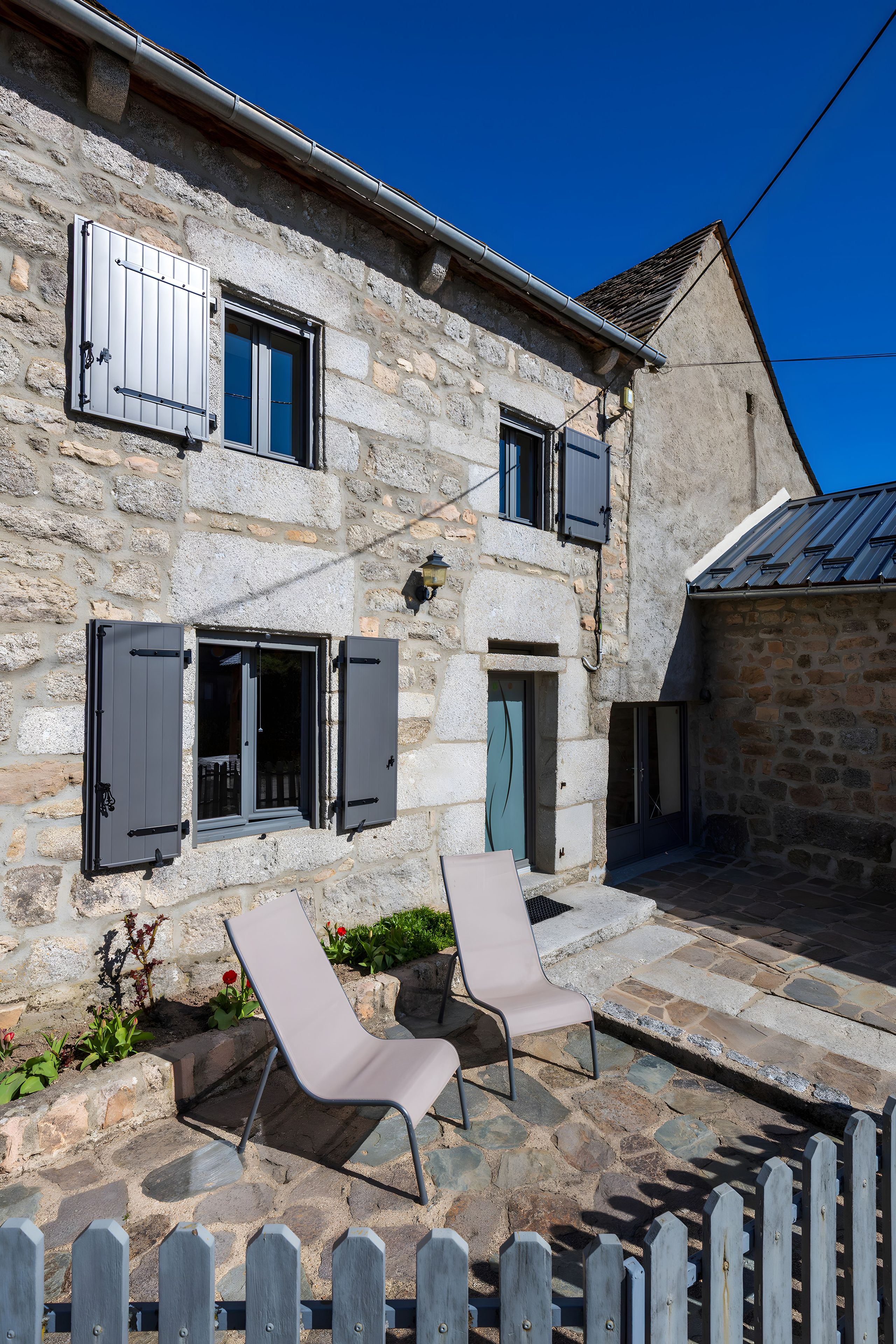 A stone building with two open windows, a door, and a small garden with two chairs.